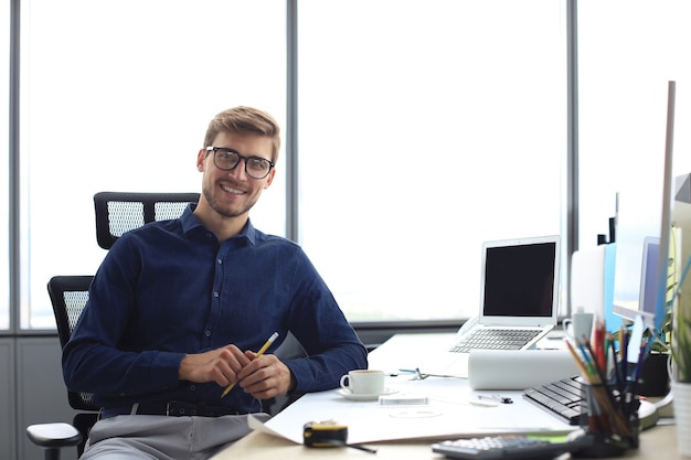 Photo bel architecte masculin regardant la caméra et souriant alors qu'il était assis au bureau.