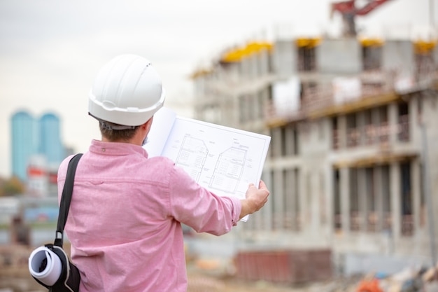 Bel architecte ou superviseur debout à l'extérieur sur un chantier de construction tenant un plan dans ses mains