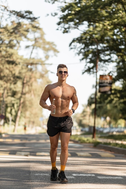 Bel athlète avec un torse nu court le long de la route dans le parc et tient une bouteille d'eau Mode de vie sain et concept sportif