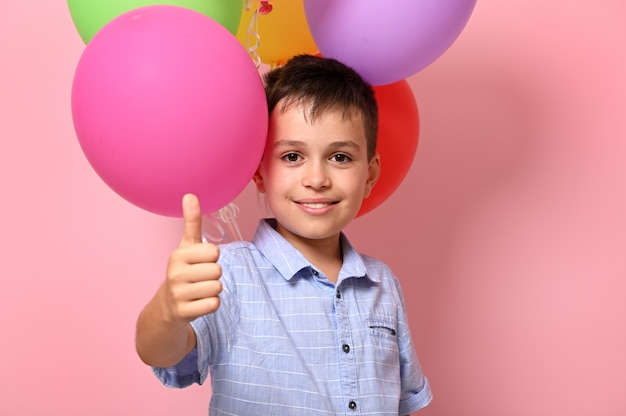 Bel écolier montrant le pouce à la caméra tout en posant sur fond rose avec des ballons colorés multicolores. Concepts. Enfance, concepts d'événements