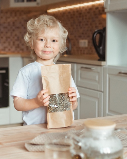 Un bel enfant heureux tient un paquet avec du thé en vrac dans ses mains à la table de la cuisine