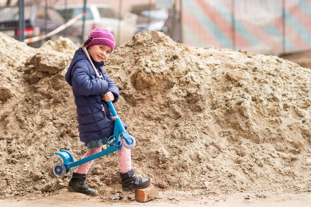 bel enfant porte un scooter sur son épaule, enjambant une pierre près d'un toboggan de sable