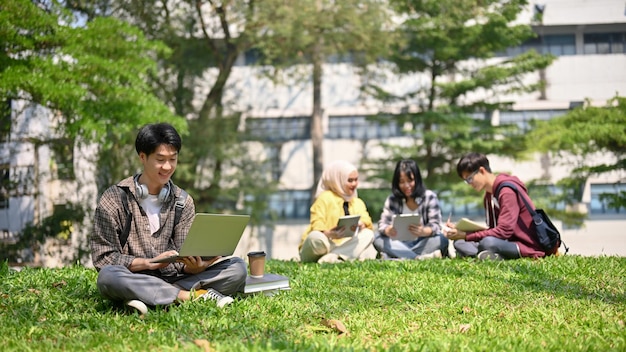 Un bel étudiant asiatique est assis sur l'herbe dans le parc du campus et utilise son ordinateur portable