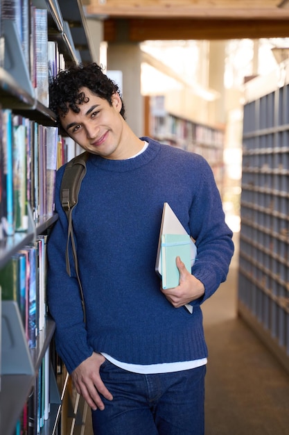 Un bel étudiant ethnique sourit à la caméra tout en choisissant des livres sur le campus de la bibliothèque universitaire