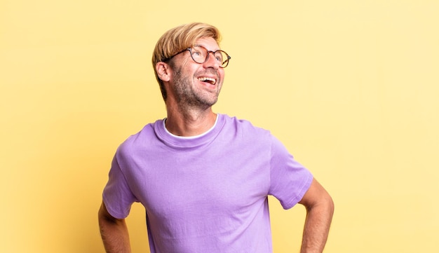 Bel homme adulte blond à l'air heureux, joyeux et confiant, souriant fièrement et regardant de côté avec les deux mains sur les hanches