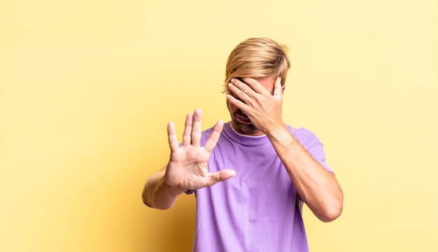 Bel Homme Adulte Blond Couvrant Le Visage Avec La Main Et Mettant L'autre Main Devant Pour Arrêter L'appareil Photo, Refusant Des Photos Ou Des Images