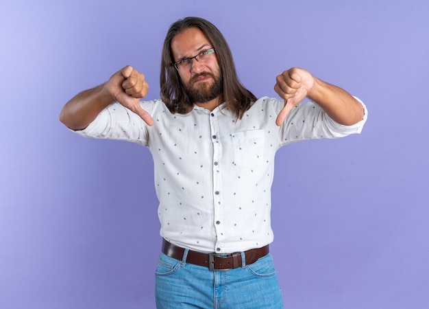 Un bel homme adulte mécontent portant des lunettes regardant la caméra montrant les pouces vers le bas isolé sur un mur violet