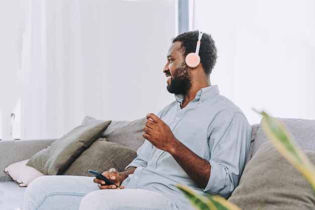 Bel homme adulte noir à la maison écoutant de la musique en ligne avec un smartphone sur un casque