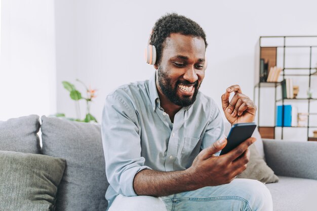 Bel homme adulte noir à la maison écoutant de la musique en ligne avec un smartphone sur un casque