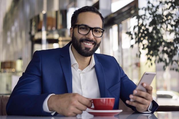 Bel homme d'affaires arabe souriant à l'aide de téléphone portable, boire du café au café