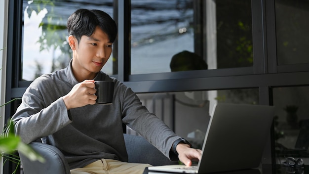 Bel homme d'affaires asiatique travaillant à distance au café en sirotant un café tout en utilisant un ordinateur portable