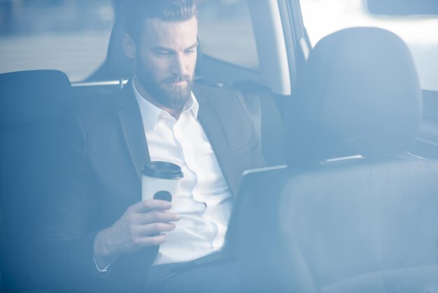 Bel homme d'affaires assis avec du café pour aller sur la banquette arrière de la voiture. Voir à travers la fenêtre