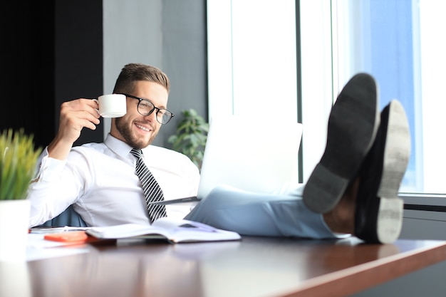 Bel homme d'affaires assis avec les jambes sur la table et buvant du café au bureau.