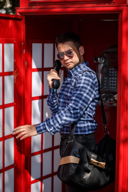 Un bel homme d'affaires avec une barbe et un sac se tient dans une cabine téléphonique rouge et parle. Beau mec en chemise et lunettes avec téléphone