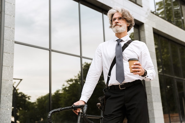 Bel homme d'affaires barbu confiant portant un costume, portant une mallette marchant à l'extérieur avec un vélo