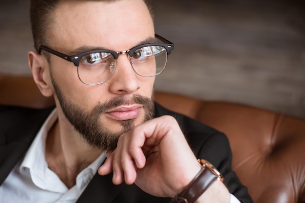 Bel homme d'affaires barbu élégant et confiant dans un costume intelligent et des lunettes assis sur un canapé au bureau