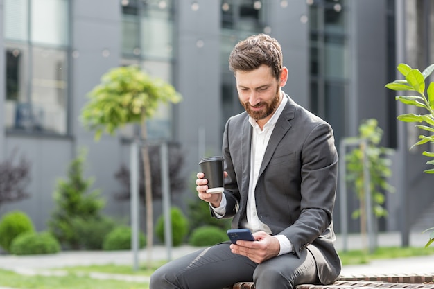 Bel homme d'affaires barbu heureux assis sur un banc avec une tasse de café utiliser un téléphone portable parcourir le smartphone dans les mains lit de bonnes nouvelles et un sourire joyeux. En dehors d'un fond de rue urbaine moderne. Parc de ville