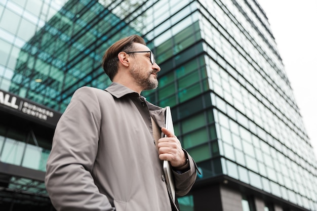 bel homme d'affaires barbu à lunettes tenant un ordinateur portable tout en se tenant près d'un immeuble de bureaux en zone urbaine