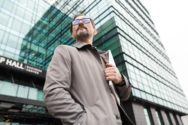bel homme d'affaires barbu à lunettes tenant un ordinateur portable tout en se tenant près d'un immeuble de bureaux en zone urbaine