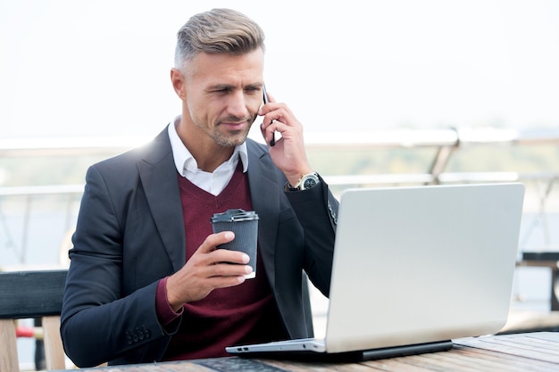 Un bel homme d'affaires boit du café en parlant au téléphone et en utilisant un ordinateur pour les affaires en ligne dans un cybercafé occupé