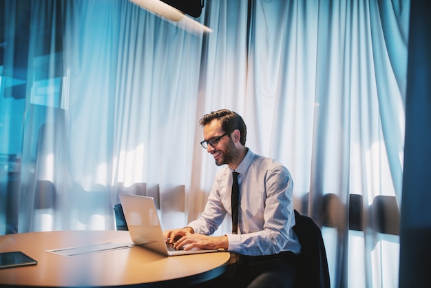 Bel homme d'affaires caucasien heureux en chemise et cravate et avec des lunettes assis au bureau et travaillant sur un projet important. Concept d'entreprise.