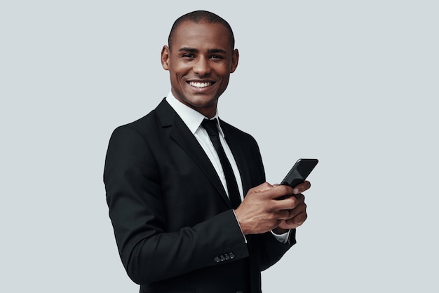 Bel homme d'affaires. Charmant jeune homme africain en tenue de soirée utilisant un téléphone intelligent et souriant en se tenant debout sur fond gris