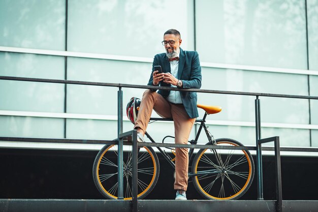 Un bel homme d'affaires décontracté d'âge moyen se rend au bureau à vélo. Il est assis sur le vélo et utilise un téléphone intelligent.