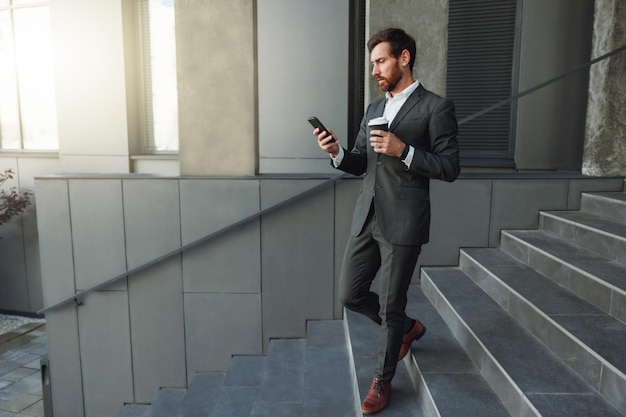 Un bel homme d'affaires descend les escaliers avec un café et un téléphone pendant une pause près du bureau