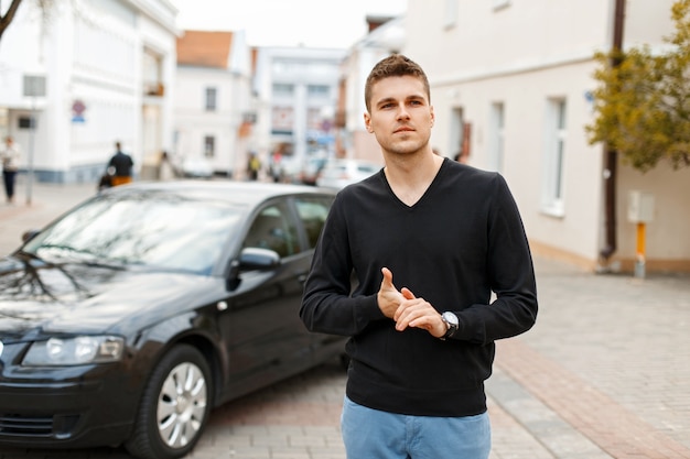 Bel homme d'affaires élégant dans un T-shirt noir et un pantalon bleu près d'une voiture noire