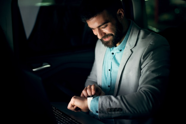 Bel homme d'affaires élégant regarde la montre sur le siège arrière d'une voiture pendant la nuit.