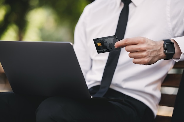 Bel homme d'affaires à l'extérieur dans le parc à l'aide d'un ordinateur portable tenant une carte de crédit.