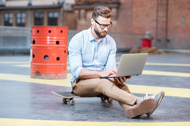 Bel homme d'affaires hipster travaillant avec un ordinateur portable assis sur la planche à roulettes sur le fond du toit. Concept d'entreprise de style de vie