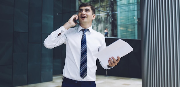 Bel homme d'affaires joyeux en vêtements formels avec des documents dans les mains parlant sur smartphone et regardant loin debout dans un immeuble de bureaux