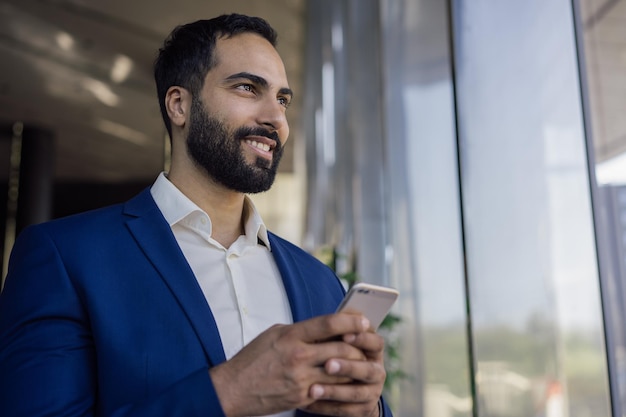 Bel homme d'affaires marocain utilisant un téléphone portable faisant des achats en ligne en regardant la fenêtre dans un bureau moderne