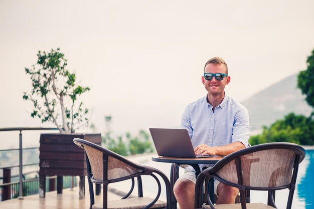 Un bel homme d'affaires masculin prospère en lunettes de soleil travaille sur un ordinateur portable assis près de la piscine Travail à distance