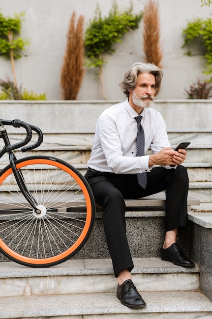 Bel homme d'affaires mature en tenue de soirée assis sur un banc avec un vélo à l'extérieur, envoyant un message par SMS