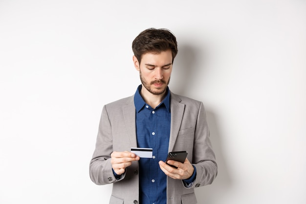 Bel homme d'affaires payant en ligne avec carte de crédit et smartphone, debout sur fond blanc en costume.