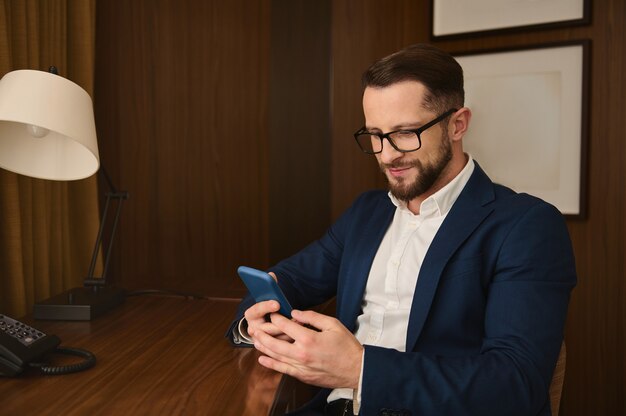 Bel homme d'affaires prospère et prospère, voyageur d'affaires, investisseur portant des lunettes, souriant, utilisant un téléphone portable assis à une table tout en se reposant dans une chambre d'hôtel pendant un voyage d'affaires