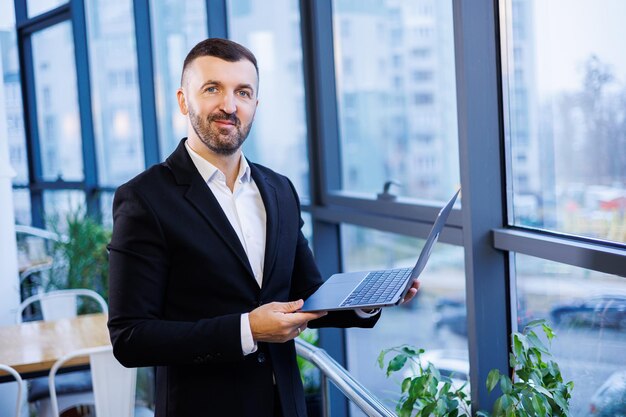 Bel homme d'affaires réussi assis devant son ordinateur portable à côté de grandes fenêtres dans un espace de bureau moderne