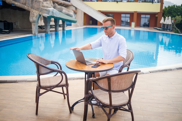 Un bel homme d'affaires réussi à lunettes de soleil travaille sur un ordinateur portable assis près de la piscine. Travail à distance. Indépendant