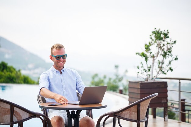 Un Bel Homme D'affaires Réussi à Lunettes De Soleil Travaille Sur Un Ordinateur Portable Assis Près De La Piscine. Travail à Distance. Indépendant