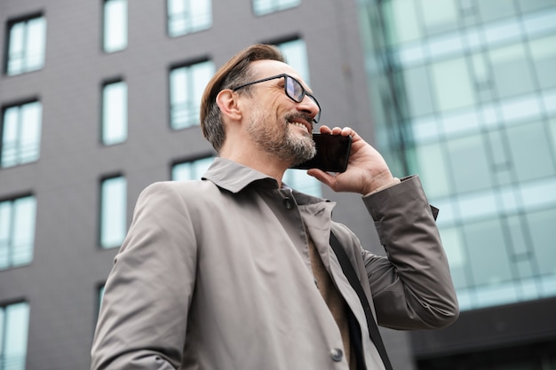 bel homme d'affaires souriant à lunettes parlant au téléphone portable tout en se tenant près d'un immeuble de bureaux en zone urbaine