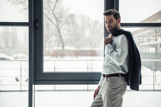 Bel homme d'affaires tient sa veste et regarde la caméra tout en se tenant près de la fenêtre du bureau