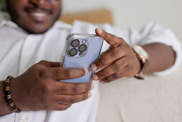 Bel homme afro-américain noir utilisant un smartphone assis sur un canapé dans un salon confortable