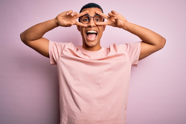 Bel homme afro-américain portant un t-shirt décontracté et des lunettes sur fond rose faisant le symbole de la paix avec les doigts sur le visage souriant joyeux montrant la victoire