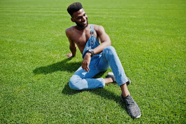 Bel homme afro-américain sexy torse nu à la salopette de jeans assis à l'herbe verte du terrain de football du stade Portrait d'homme noir à la mode