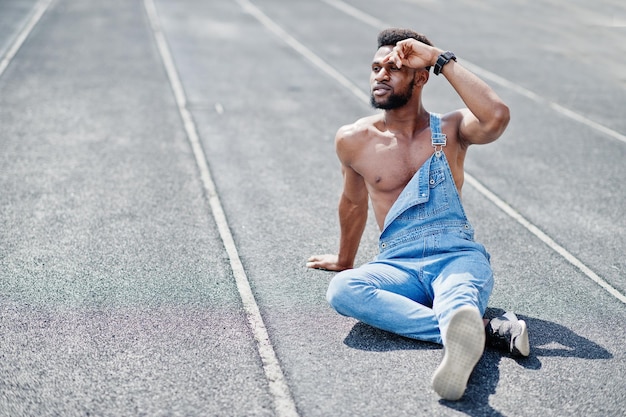 Bel homme afro-américain sexy torse nu à la salopette de jeans assis à l'hippodrome du stade Portrait d'homme noir à la mode