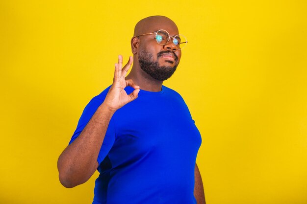 Bel homme afro-brésilien portant des lunettes chemise bleue sur fond jaune signe ok d'accord bien fait approbation bon grand