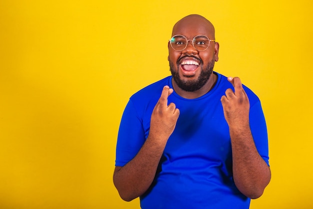 Bel homme afro-brésilien portant des lunettes chemise bleue sur fond jaune Signe porte-bonheur acclamant souhaitant vouloir chance vibrante bonne chance dessiner