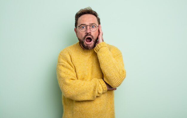 Bel homme d'âge moyen bouche bée sous le choc et l'incrédulité avec la main sur la joue et le bras croisé se sentant stupéfait et étonné